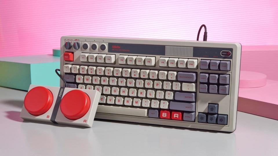 A gray and red mechanical keyboard called the 8BitDo Retro Mechanical Keyboard rests against a well-lit pink and pastel green background. In front of the keyboard are a pair of giant red 